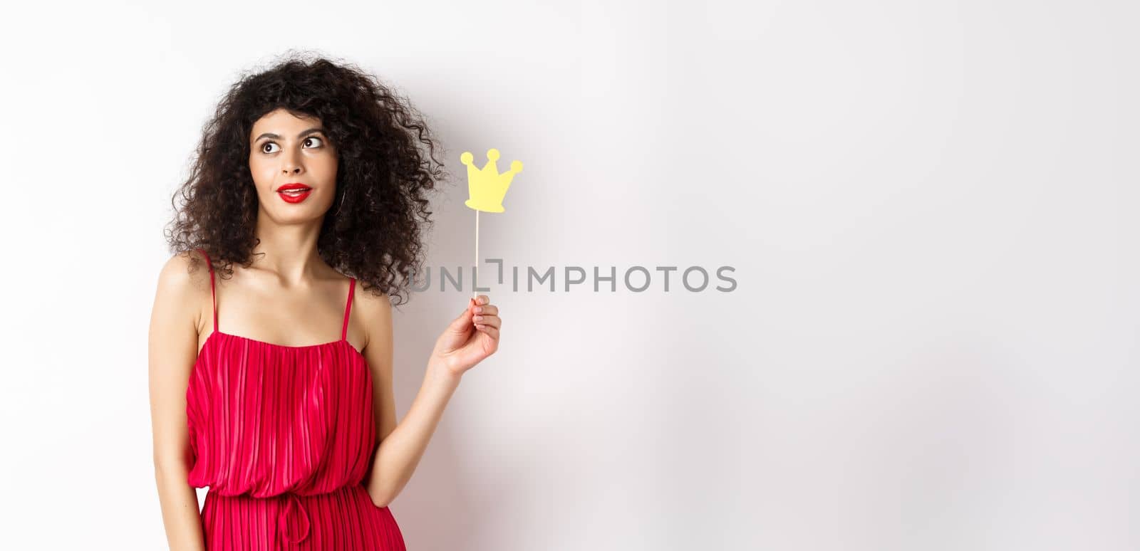 Stylish woman with curly hair in red dress, holding queen crown on stick and looking aside, standing on white background.