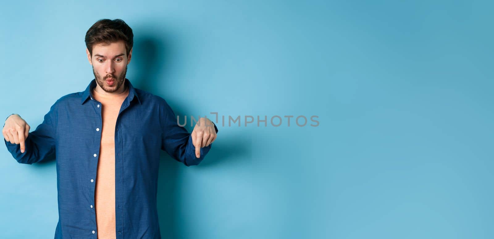 Impressed caucasian male model in casual clothes, say wow, looking and pointing fingers down at logo, standing on bllue background.