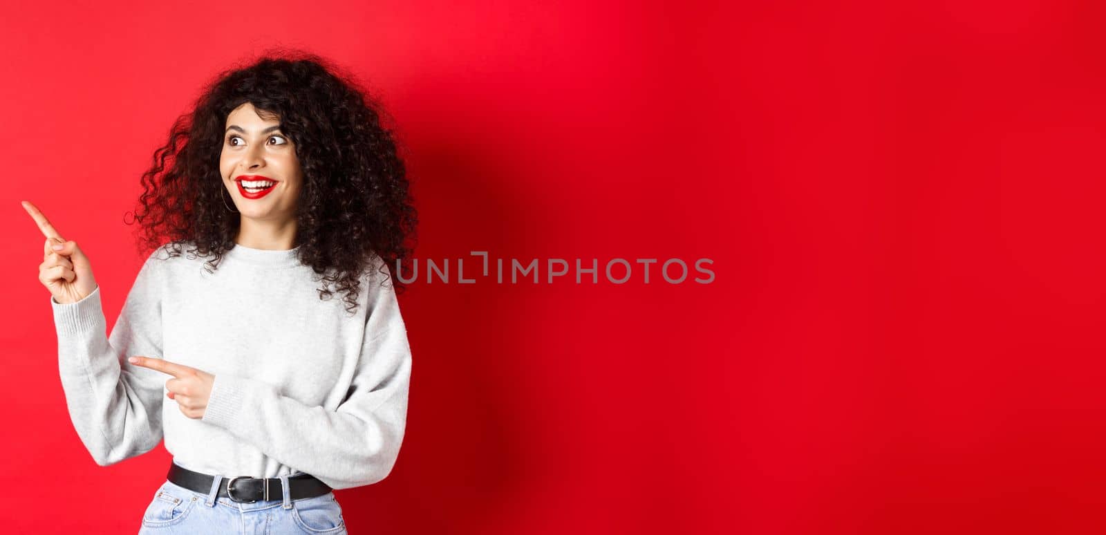 Portrait of excited curly woman with red lips, looking and pointing left at logo with amazed face, checking out promo deal, standing on studio background.