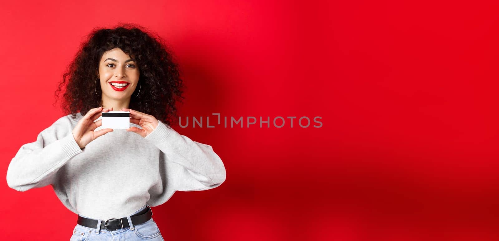 Happy lady with curly hair showing plastic credit card and smiling, standing against red background. Shopping concept.
