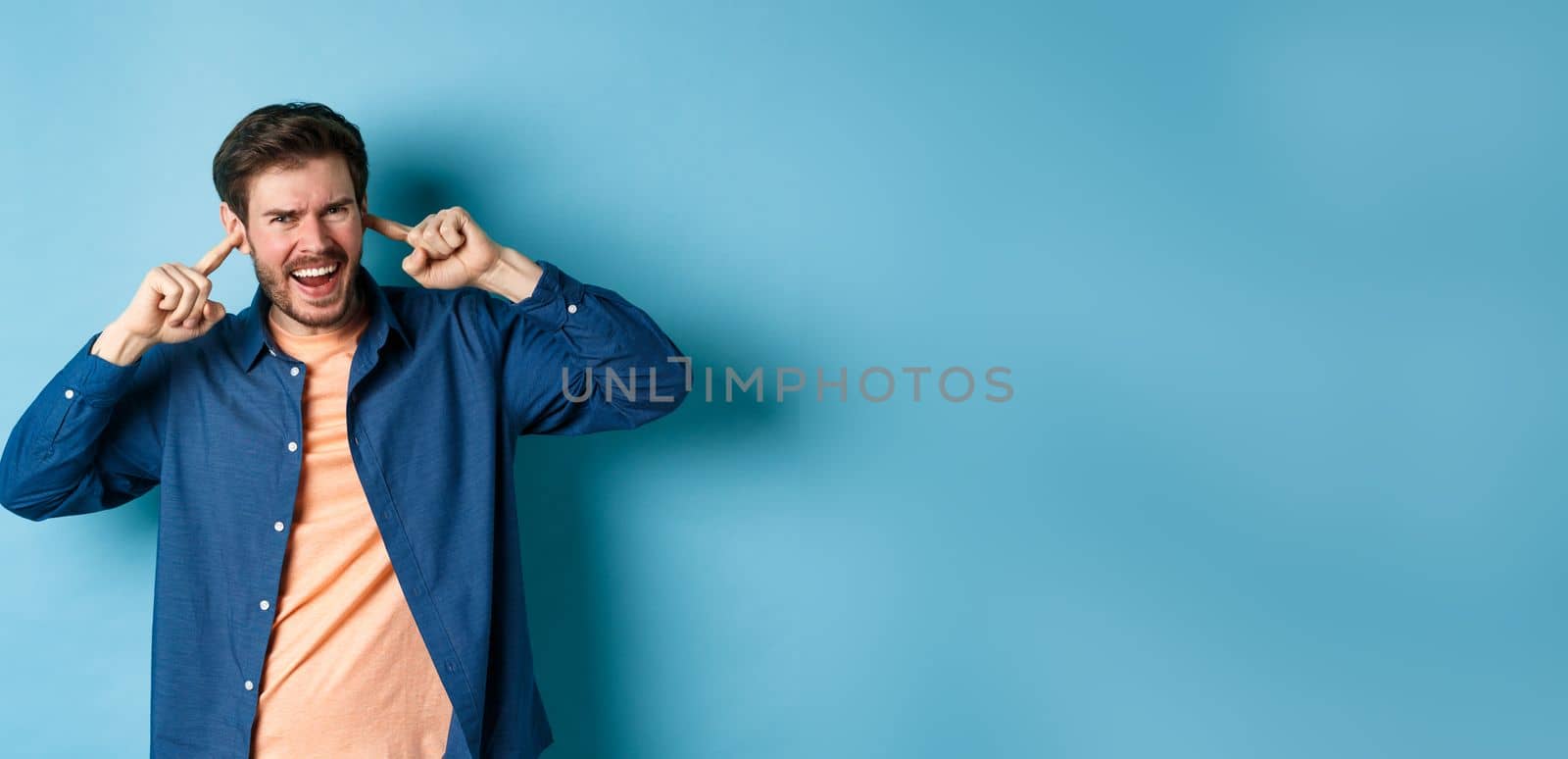 Angry guy shouting with shut ears, block loud music and frowning annoyed, standing on blue background.