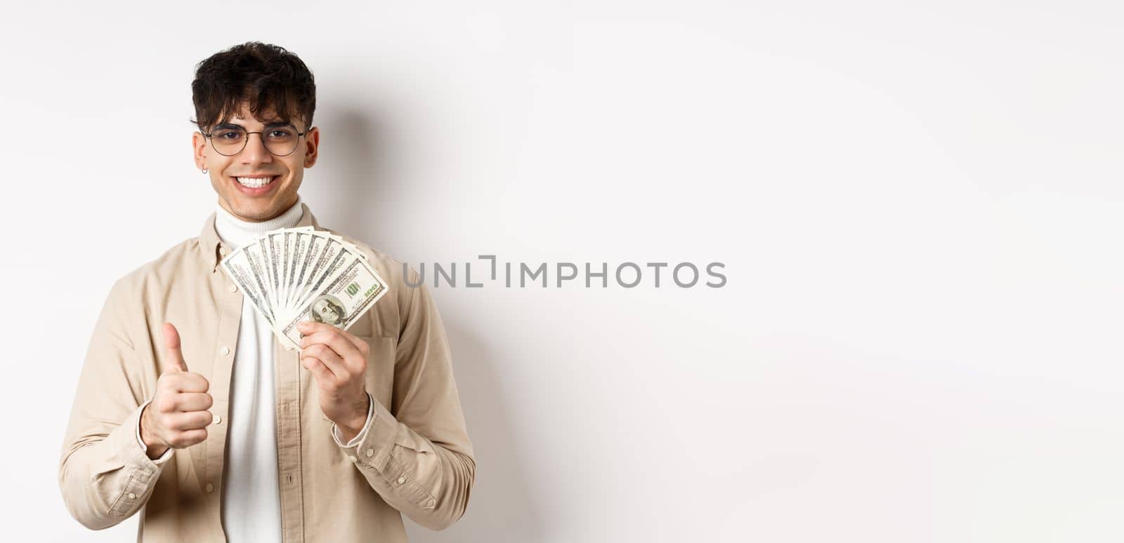 Young guy in glasses showing dollar bills and thumbs up, making money, standing with cash on white background.