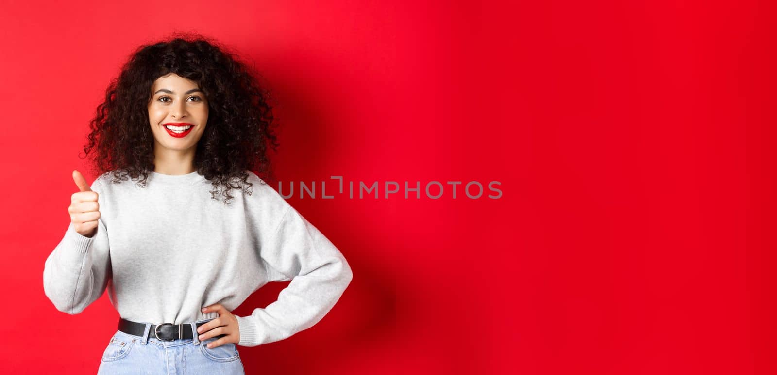 Enthusiastic young woman with curly hair, red lips, showing thumbs up and smiling in approval, praise good product, standing against red background.
