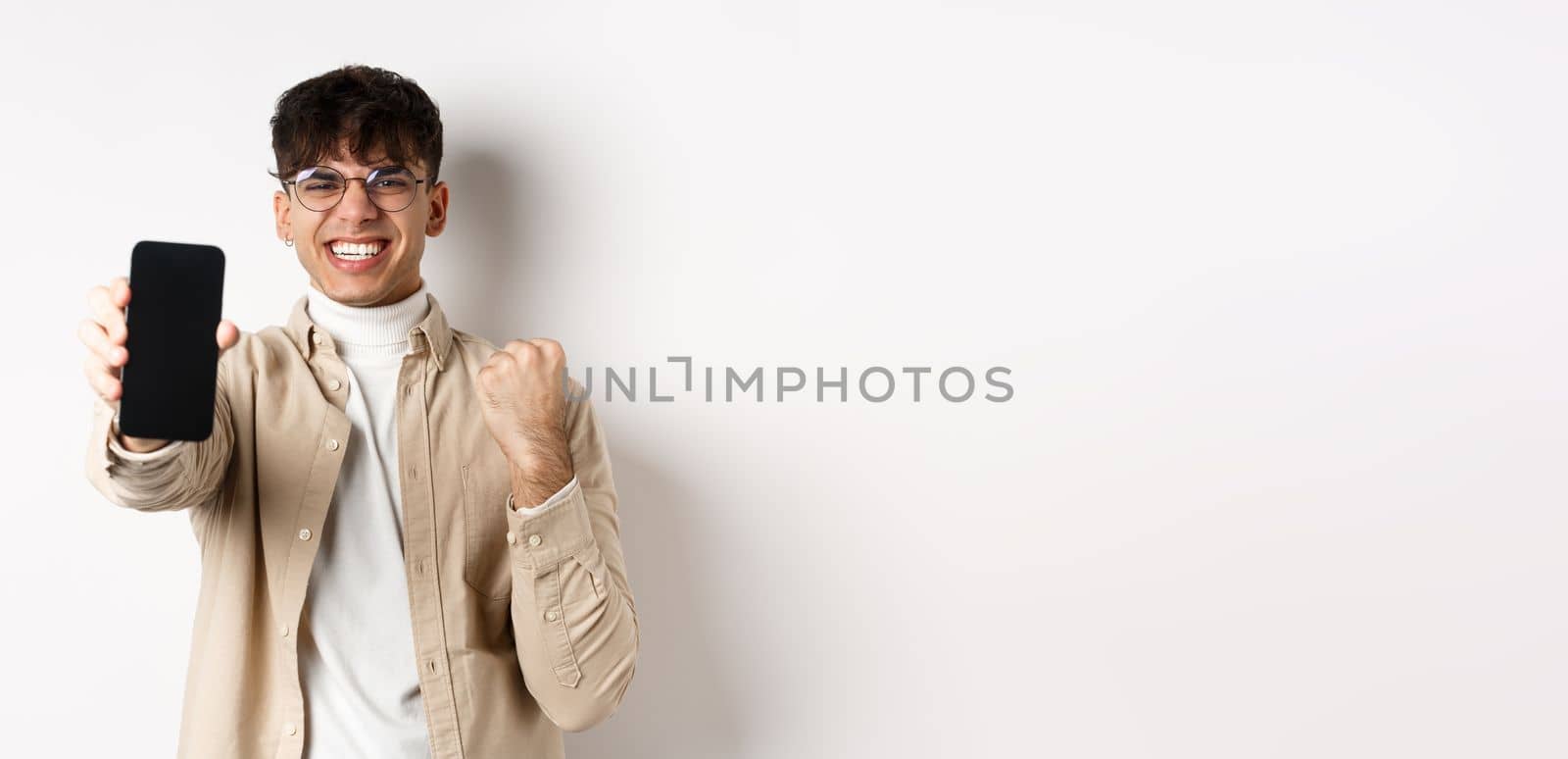 Lucky young man winning prize online, showing smartphone screen and rejoicing of good news, standing happy on white background by Benzoix
