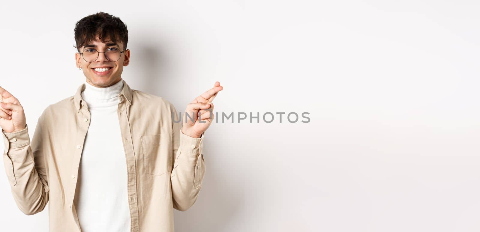 Handsome and positive young man smiling, making wish with crossed fingers and happy face, hope for dream to come true, standing on white background by Benzoix