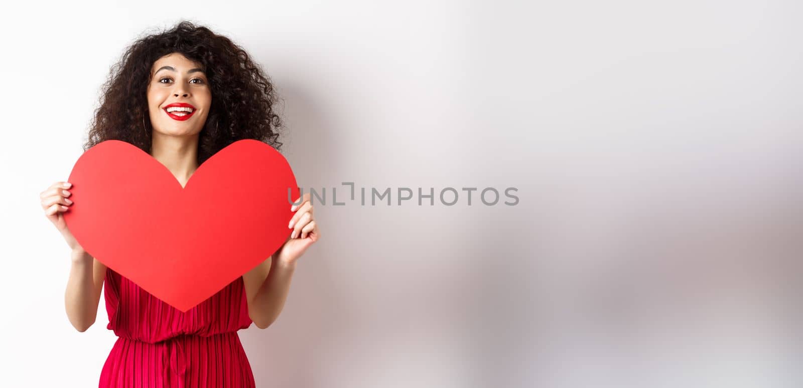 Romantic woman in dress showing big red heart, falling in love, smiling happy at camera, white background by Benzoix