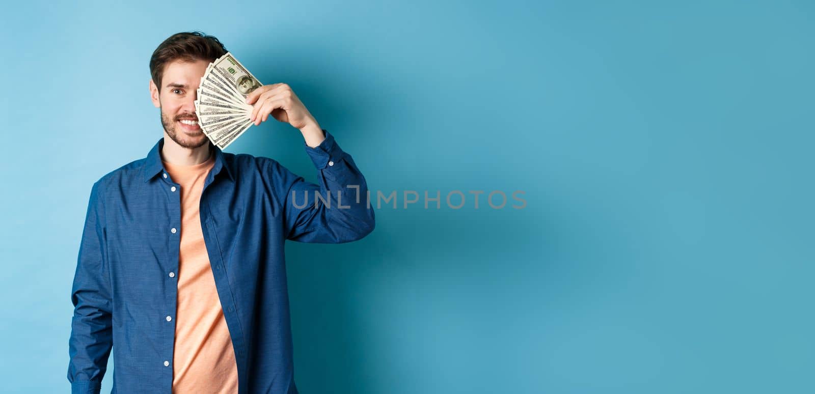 Happy young guy covering half of face with dollars and smiling, winning prize money, standing on blue background.