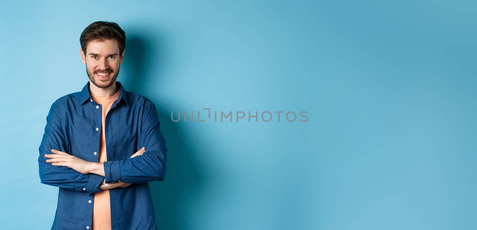 Confident young man feeling like professional, cross arms on chest and smiling sassy, standing on blue background.