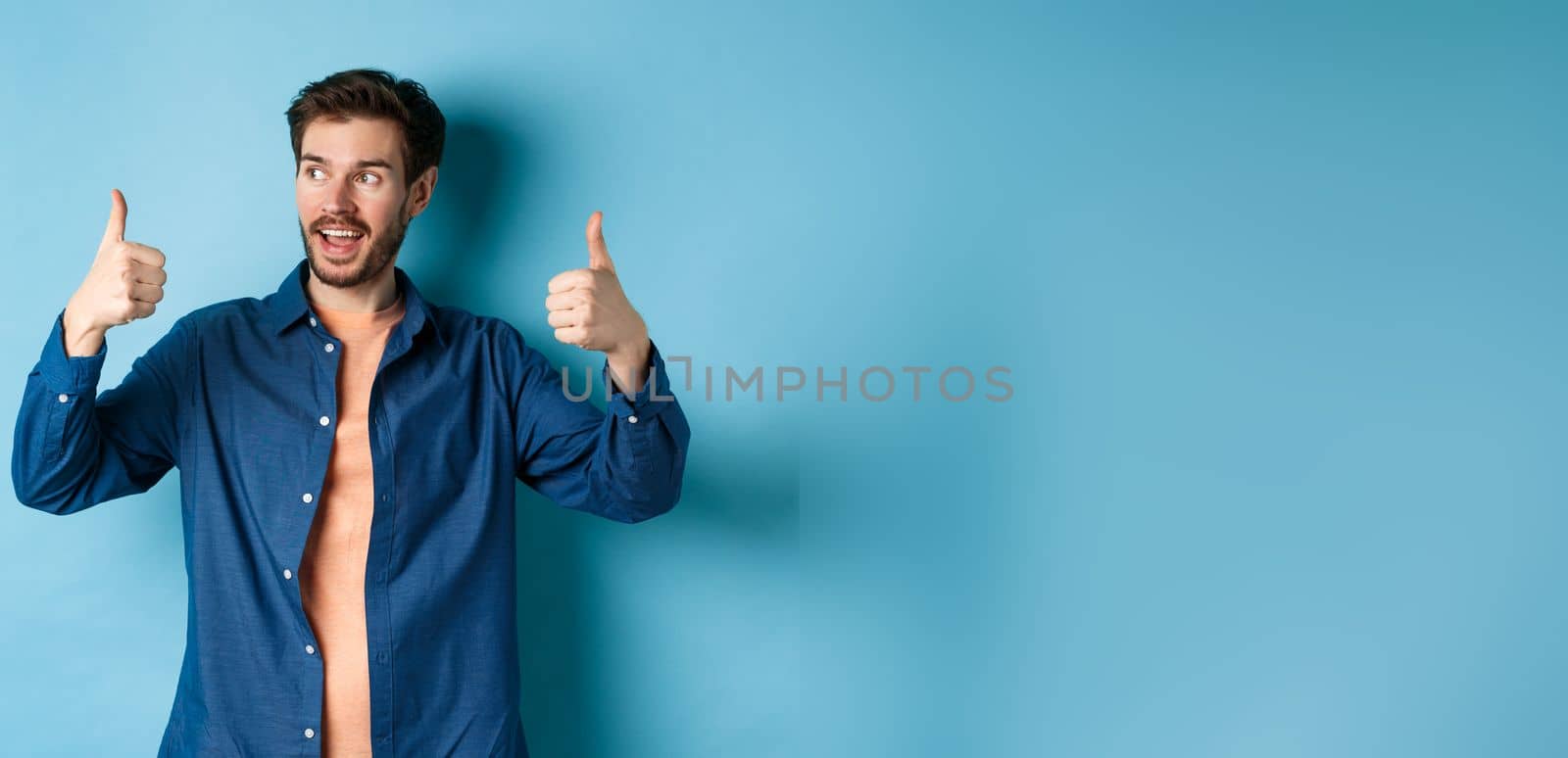 Handsome young man looking aside at empty space and showing thumbs up, standing on blue background by Benzoix