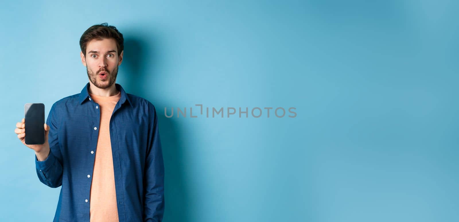 Impressed caucasian male model showing smartphone screen and look amazed, talking about online offer, standing on blue background by Benzoix