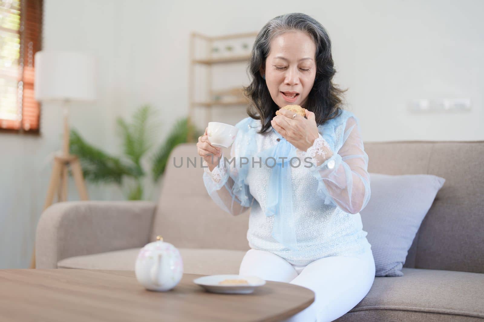 Portrait of an elderly Asian woman drinking healthy tea while eating snack. by Manastrong