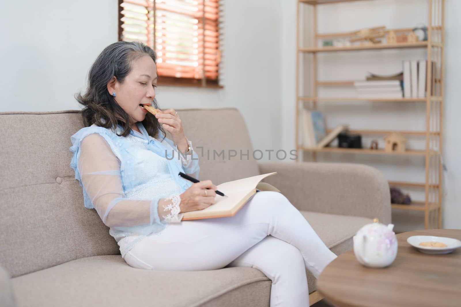 Portrait of an elderly Asian woman holding a notebook eating snacks and drinking tea. by Manastrong
