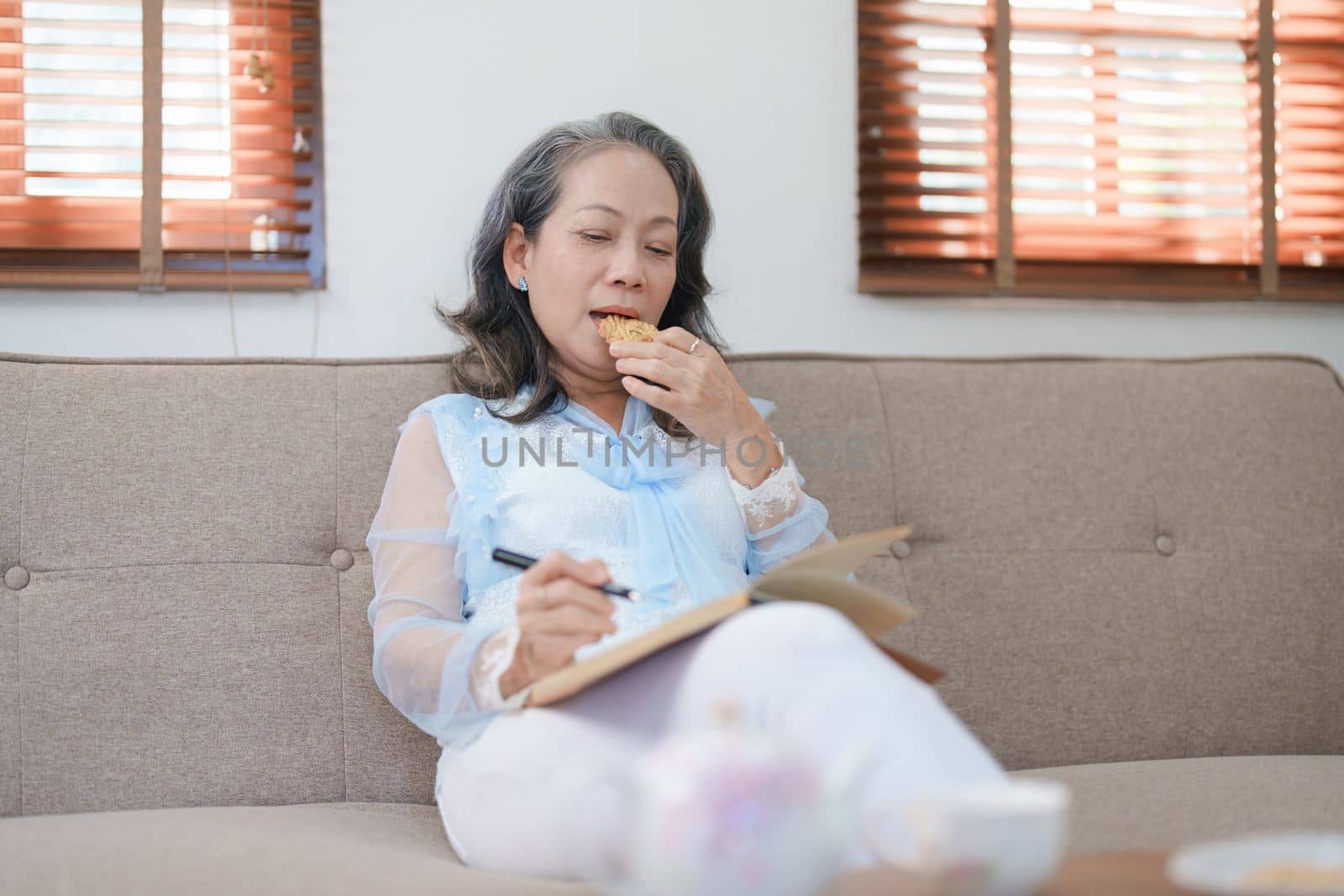 Portrait of an elderly Asian woman holding a notebook eating snacks and drinking tea