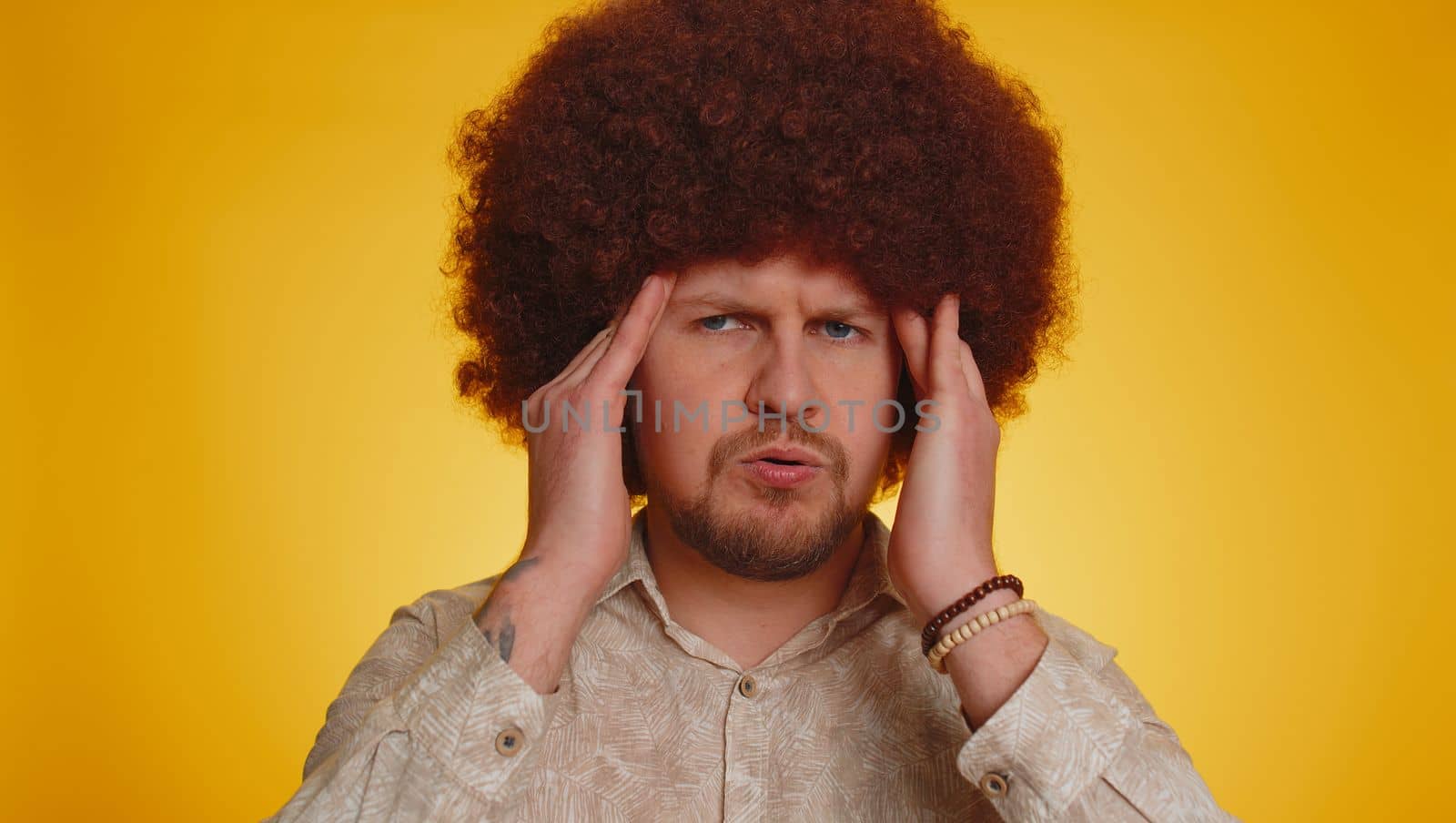 Bearded young man with lush afro hairstyle coiffure rubbing temples to cure headache problem, suffering from tension and migaine, stress, grimacing in pain high blood pressure on yellow background