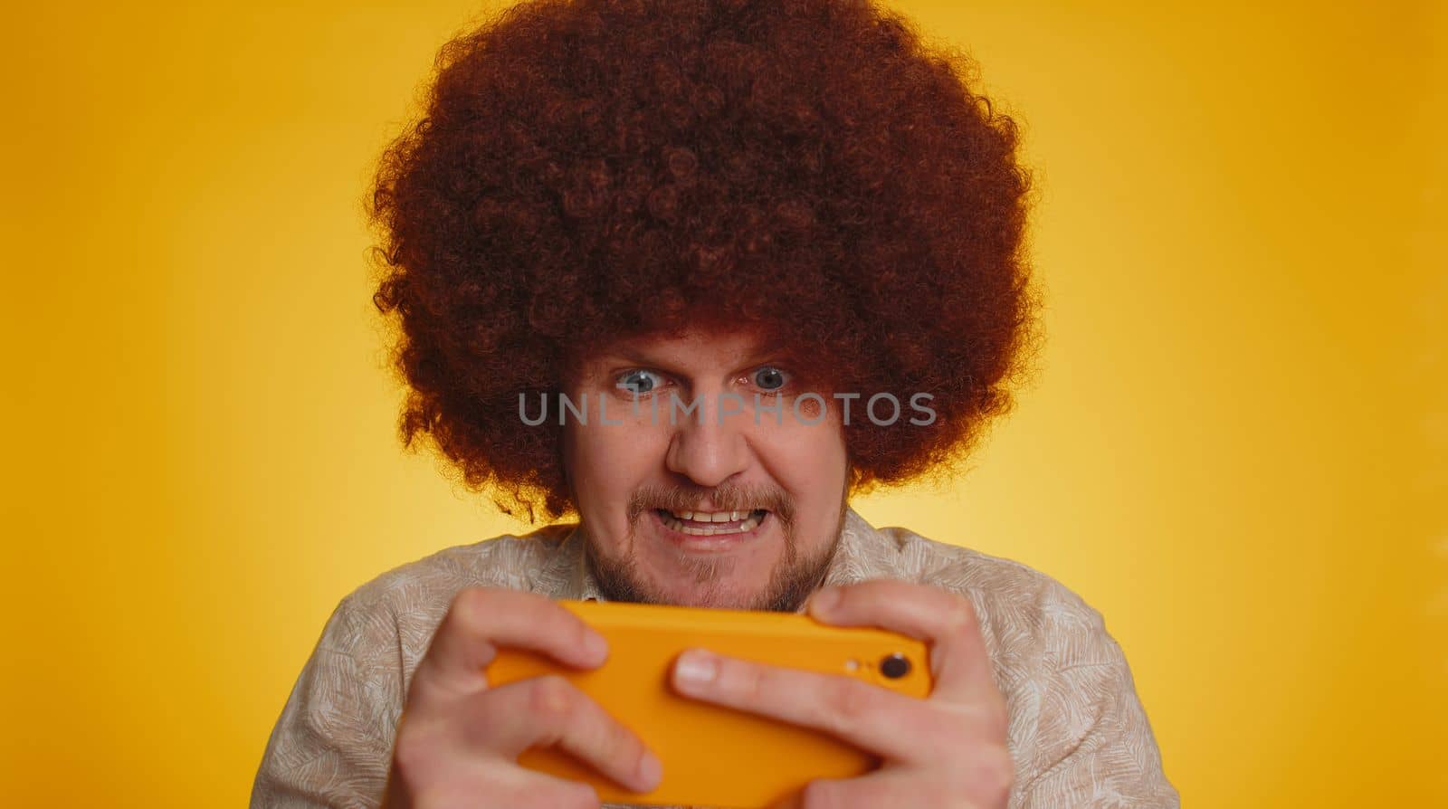 Worried bearded man with afro hairstyle coiffure enthusiastically play racing or shooter video games on smartphone. Hipster guy use mobile phone gadget app with drive simulator on yellow background