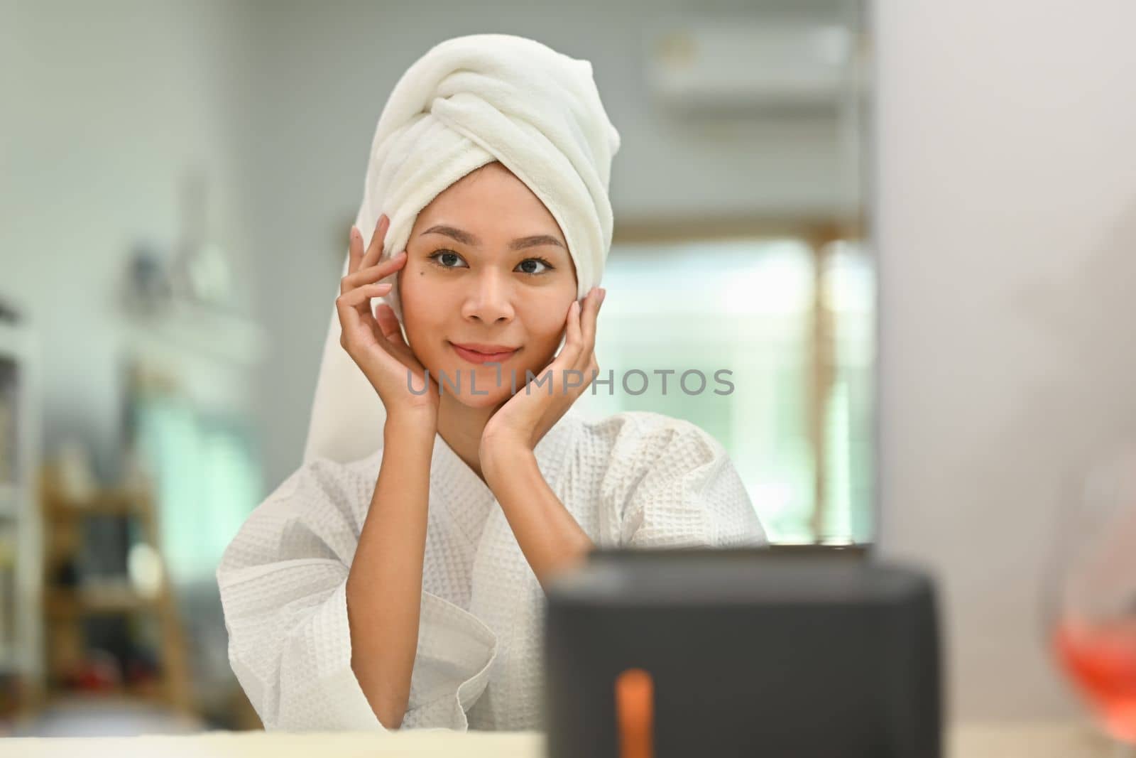 Gorgeous woman with towel on head touching face and looking at mirror. Home care and beauty treatment concept by prathanchorruangsak