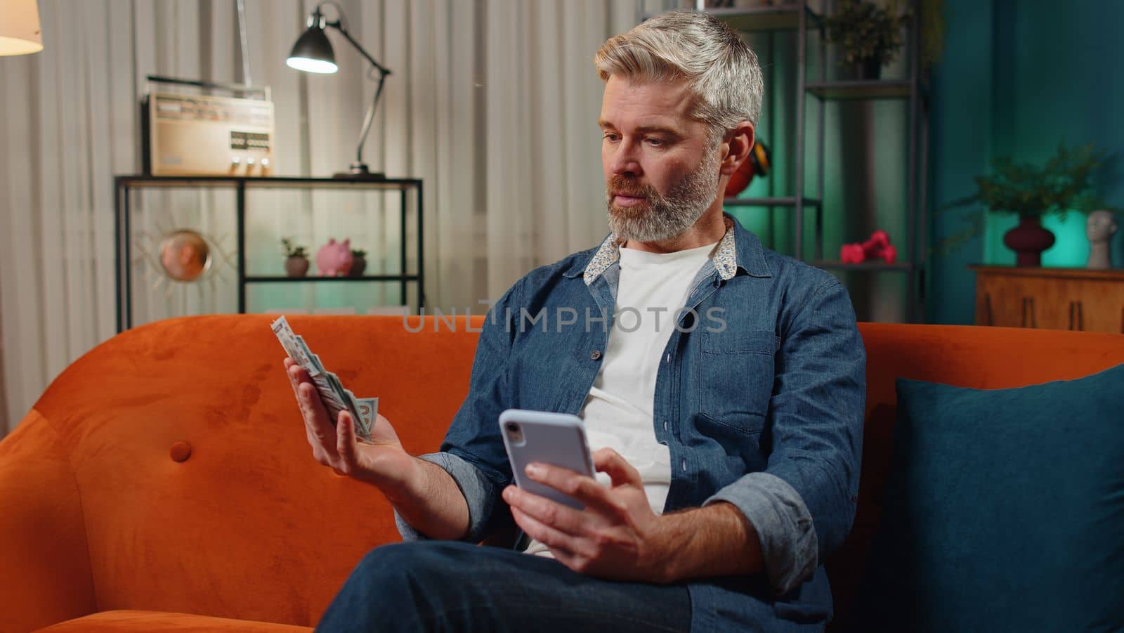 Planning family budget. Bearded senior man counting money dollar cash use smartphone calculate domestic bills at home. Joyful middle-aged guy satisfied of income saves money for planned vacation gifts