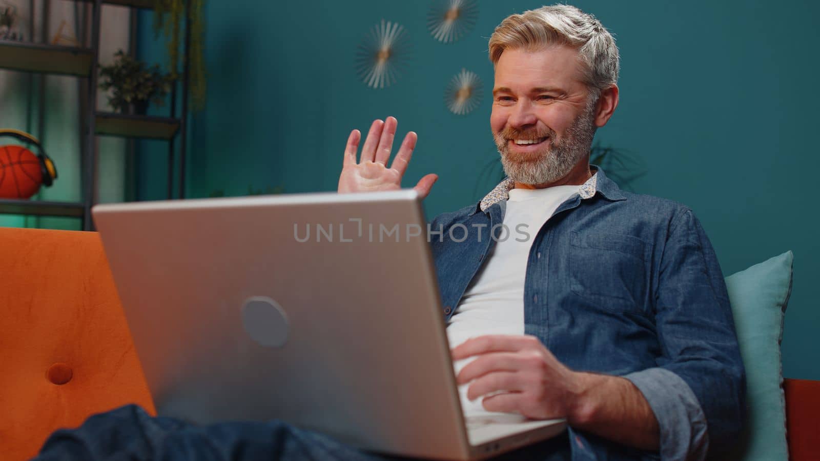 Middle-aged senior man sitting on couch looking at camera, making video webcam conference call with friends or family, enjoying pleasant conversation. Mature guy laughing waving hello at night home