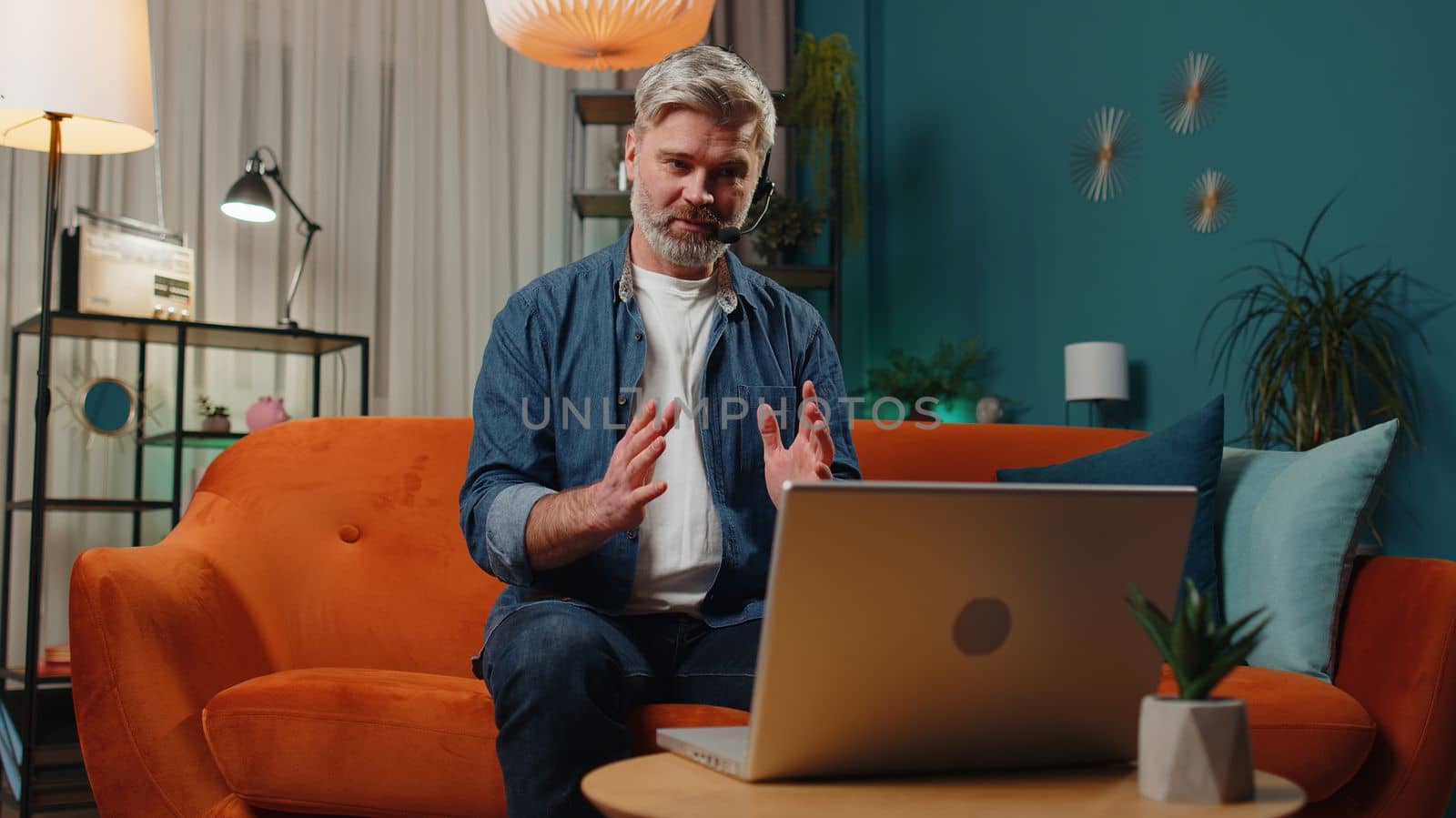 Senior man at home wearing headset, freelance worker, call center or support service operator helpline, having talk with client or colleague communication support. Mature old guy in night evening room