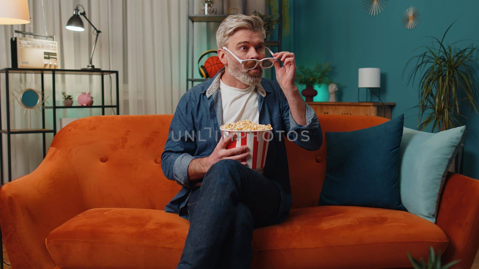 Excited senior mature man sits on couch eating popcorn snacks and watching interesting TV serial sport game, film, online social media movie content at home. Elderly guy enjoying evening entertainment