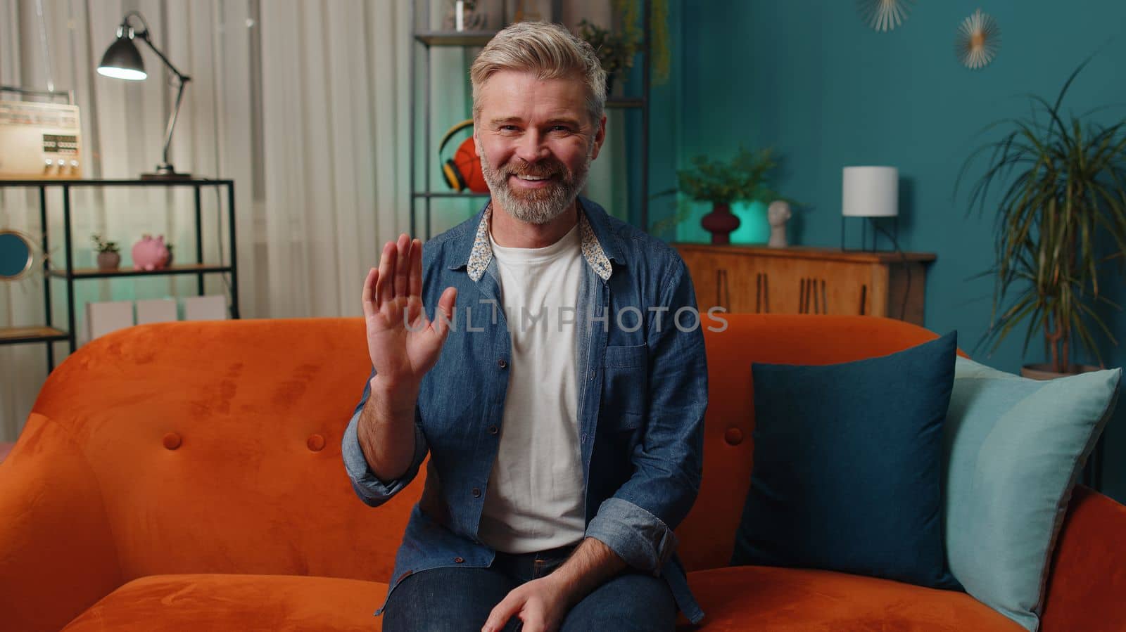 Middle-aged man smiling friendly at camera and waving hands gesturing hello, hi, greeting or goodbye, welcoming with hospitable expression. Mature old gray-haired guy indoors at home in room on couch
