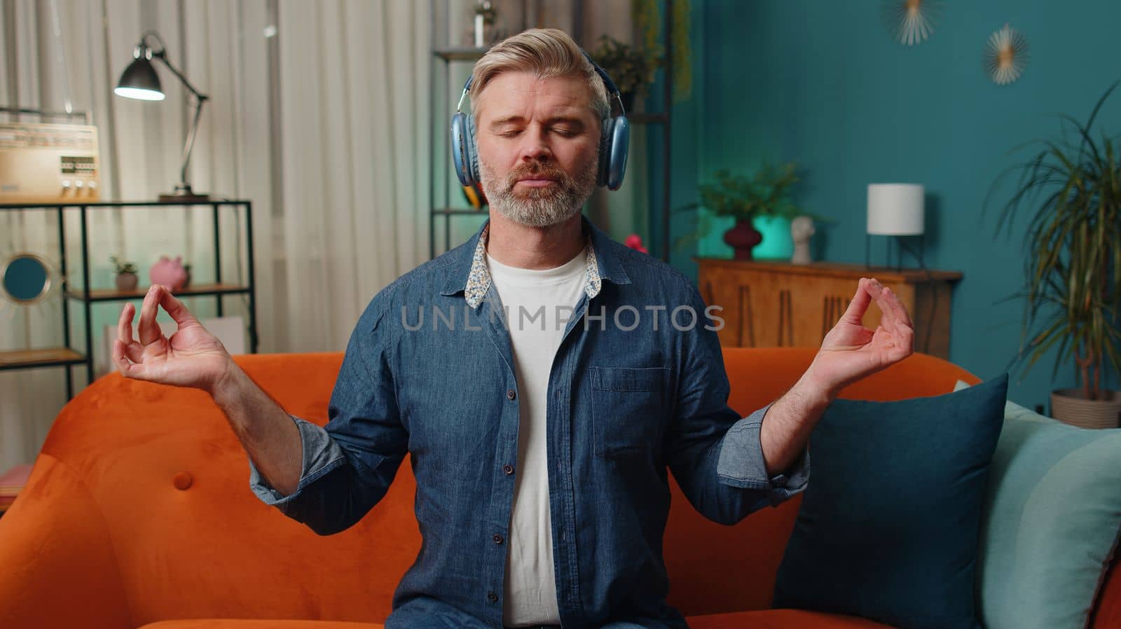 Keep calm down, relax. Middle-aged old man at home couch breathes deeply with mudra gesture, eyes closed meditating with concentrated thoughts, peaceful mind, listen music. Mature guy in evening room