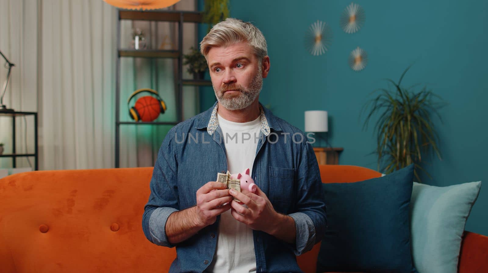 Poor middle-aged man insufficient amount of money, holding piggybank and one dollar banknote at home room. Financial crisis. Bankruptcy. Poverty and destitution at home. Mature senior gray-haired guy