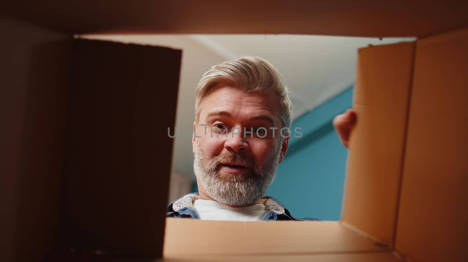 Happy surprised winner man unpacking delivery parcel at home. Smiling satisfied senior guy shopper online shop customer opening cardboard box receiving purchase gift headphones by fast postal shipping