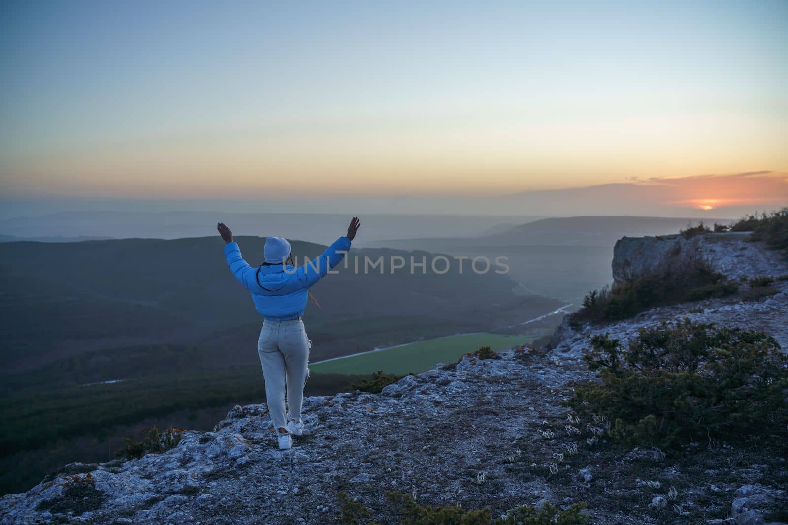 Woman hiker open arms on top of sunrise mountain. The girl salutes the sun, wearing a blue jacket, white hat and white jeans. Conceptual design. by Matiunina