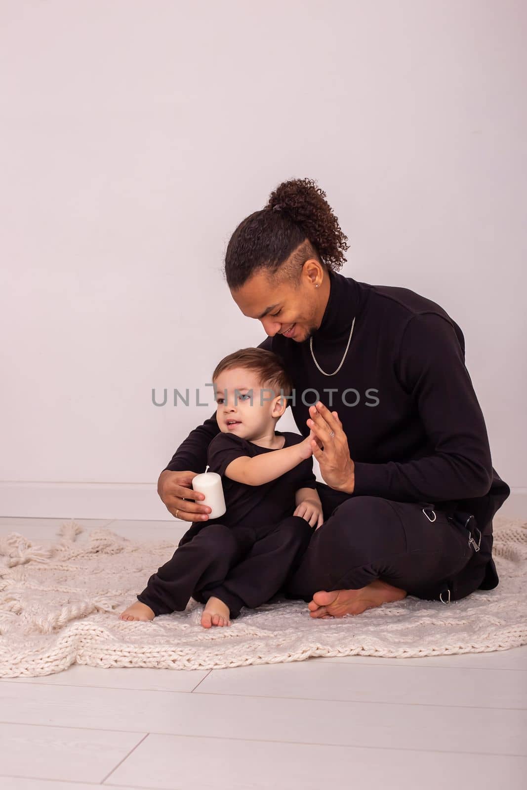 Stylish father and son, in black clothes, sitting on a knitted blanket, on the floor in the room, light interior, having fun playing, holding a large white candle. Vertical. Copy space