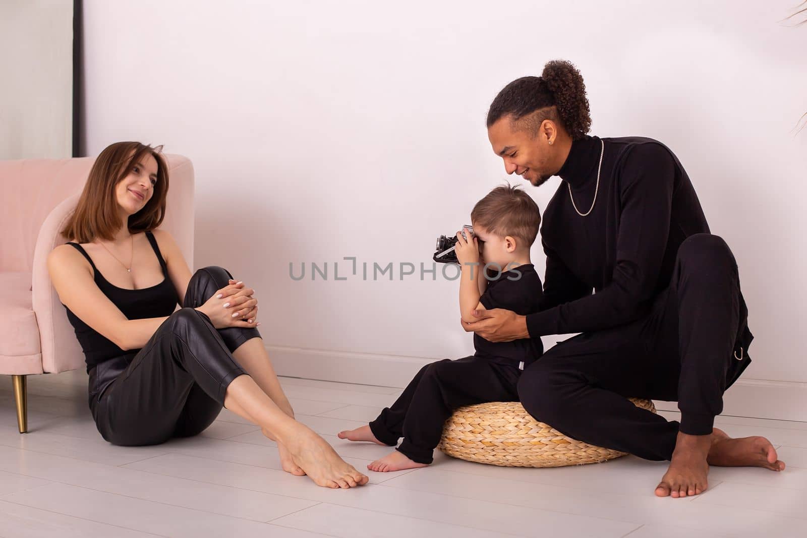 A happy multiracial family in black clothes, with a little boy with an old camera, taking pictures of his mother, in a bright living room. Copy space