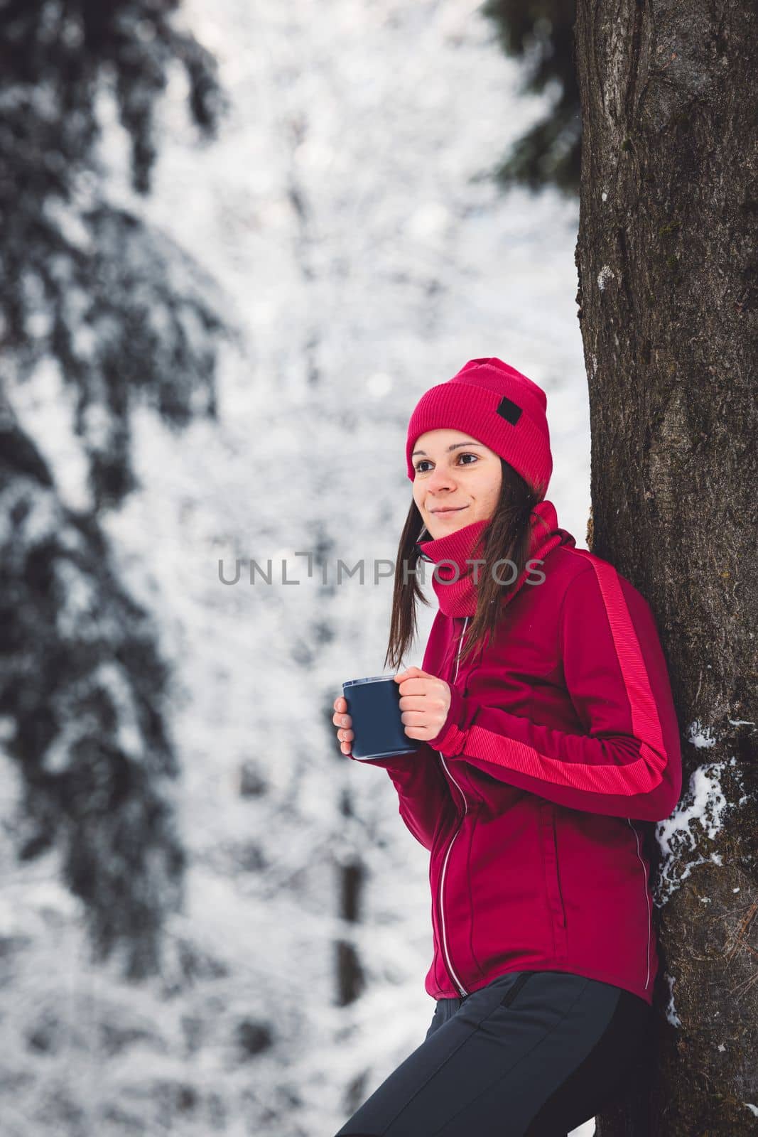 Beautiful young caucasian woman with brown hair, wearing a red jacket and a hat outside in the forest when it's snowing. Woman walking around a snowy forest. Woman holding a to go cup with tea.