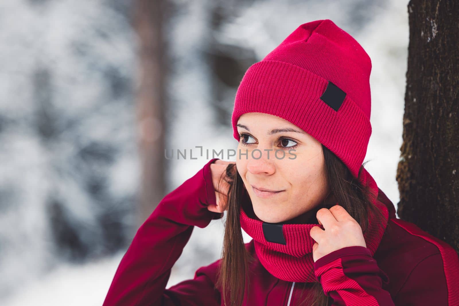 Beautiful young caucasian woman with brown hair, wearing a red jacket and a hat outside in the forest when it's snowing. Woman walking around a snowy forest. Woman holding a to go cup with tea.