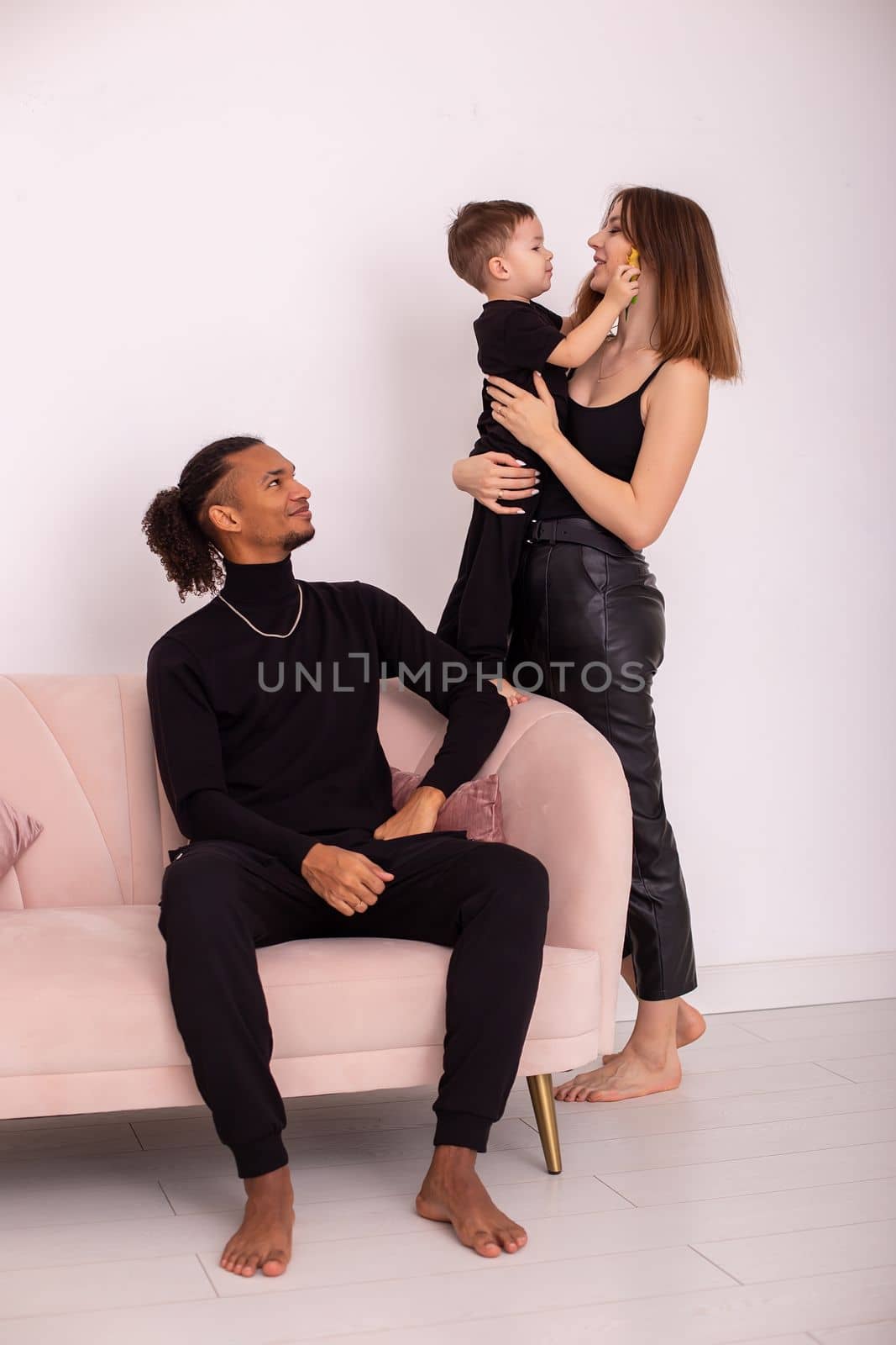 Happy multiracial family with little boy, in black clothes, with bare foot , sits in the light living room on light pink sofa. Vertical. Copy space