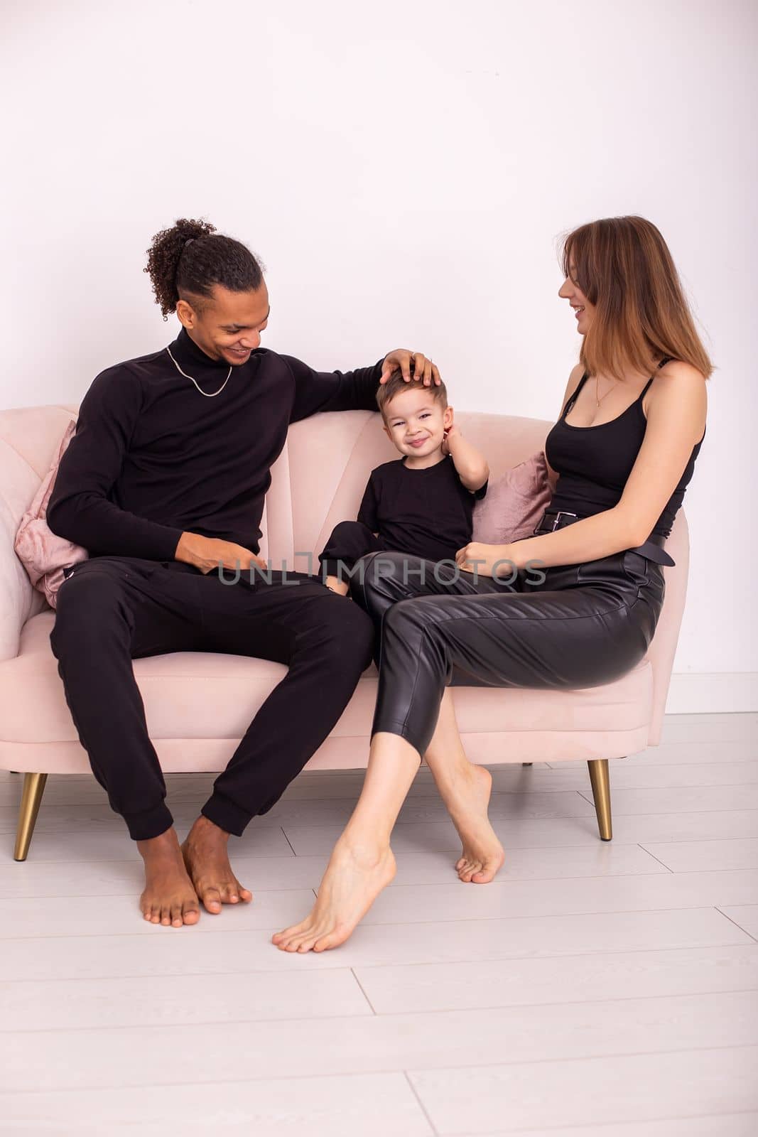 Beautiful multiracial family with little boy, in black clothes, with bare foot , sits in the light living room on light pink sofa, communicate. Vertical. Copy space