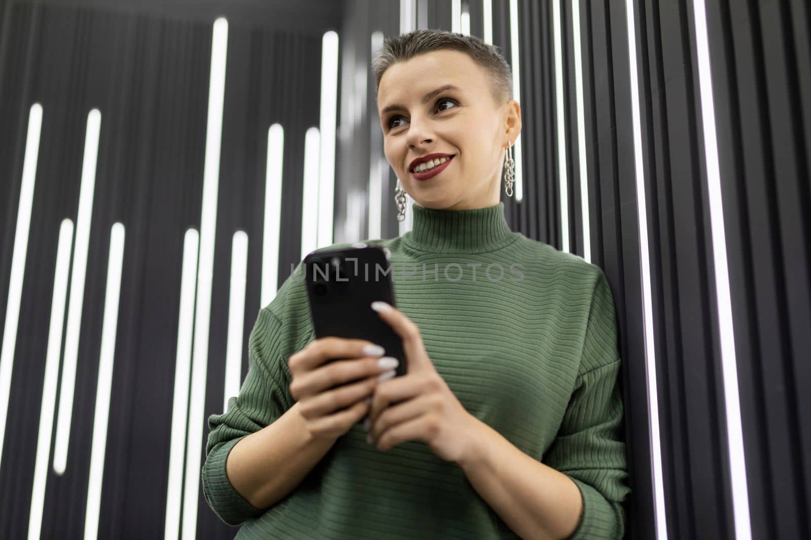 attractive caucasian woman with short haircut with mobile phone near gray backlit wall.