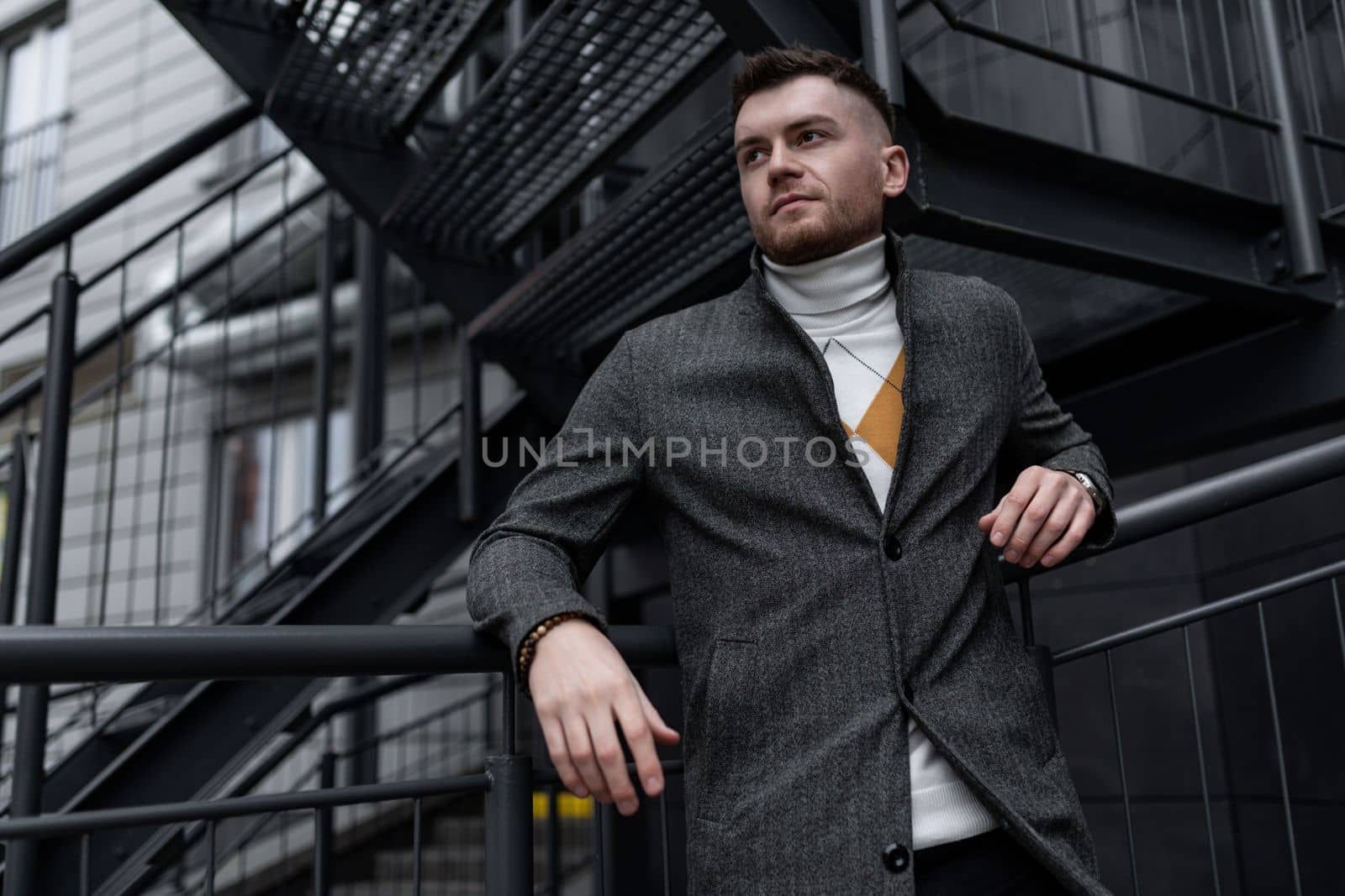 a strong young man in a gray coat stands on the metal stairs of a trendy building.