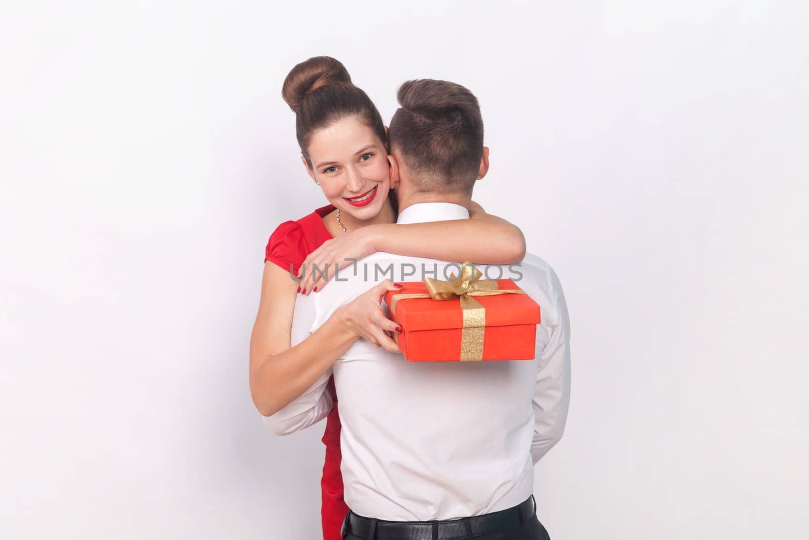 Smiling woman and man in elegant clothes standing together, guy posing backwards, his wife hugging him and holding present box. Indoor studio shot isolated on gray background.
