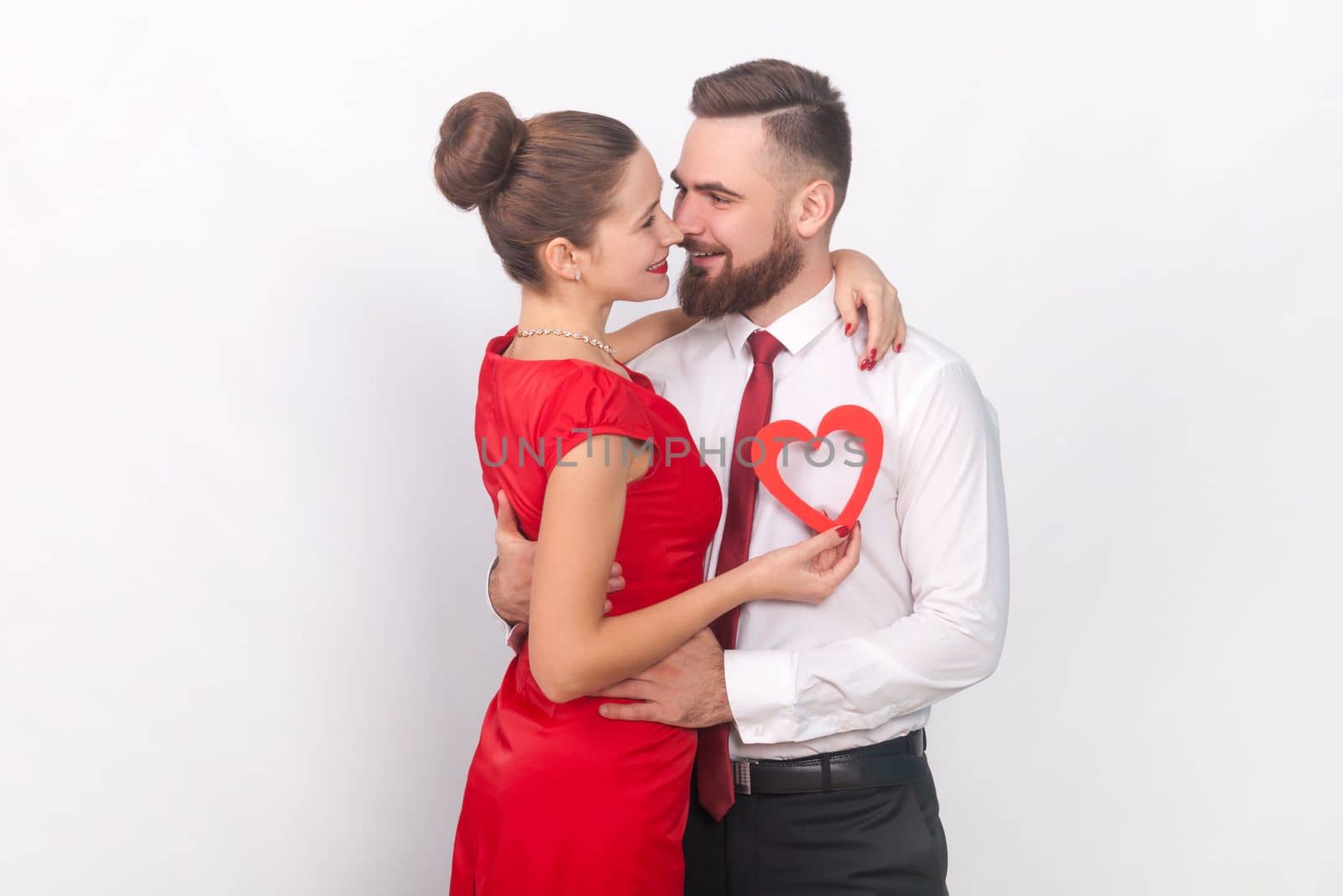 Romantic loving woman and man in elegant clothes standing together, hugging each other, showing little red heart, celebrating anniversary. Indoor studio shot isolated on gray background.