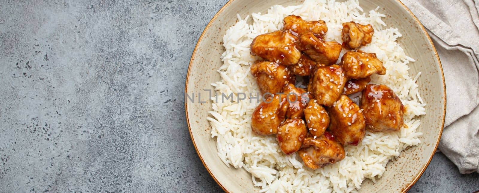 Chinese sweet and sour sticky chicken with sesame seeds and rice on ceramic plate with chopsticks top view on gray rustic stone background, traditional dish of China. Space for text