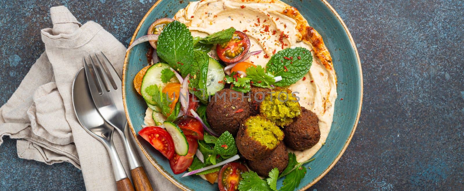 Middle Eastern Arab meal with fried falafel, hummus, vegetables salad with fresh green cilantro and mint leaves in ceramic bowl on stone rustic background top view. Arabian style breakfast or dinner