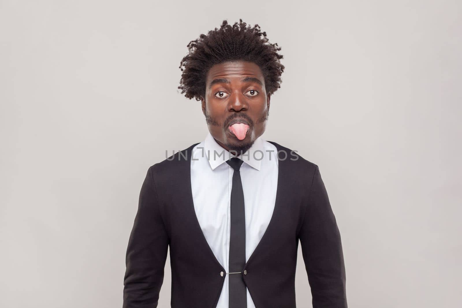 Portrait of childish man with Afro hairstyle sticking out tongue, expressing her dislike or disregard towards something, wearing white shirt and tuxedo. Indoor studio shot isolated on gray background.