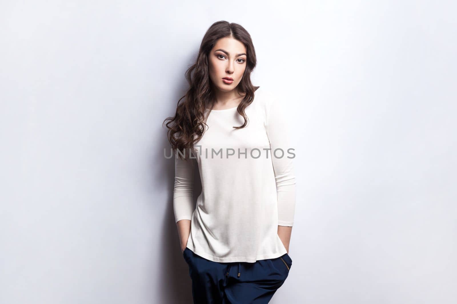 Portrait of calm serious woman with wavy hair wearing white shirt standing with hands in pockets, looking at camera with strict confident expression. Indoor studio shot isolated on gray background.