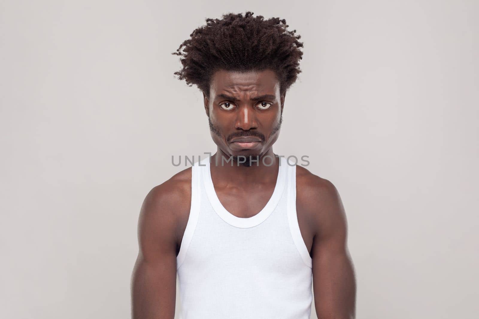 Sad unhappy man with Afro hairstyle frowning face, being upset with bad news he received, looking at camera with pout lips, wearing white T-shirt. Indoor studio shot isolated on gray background.