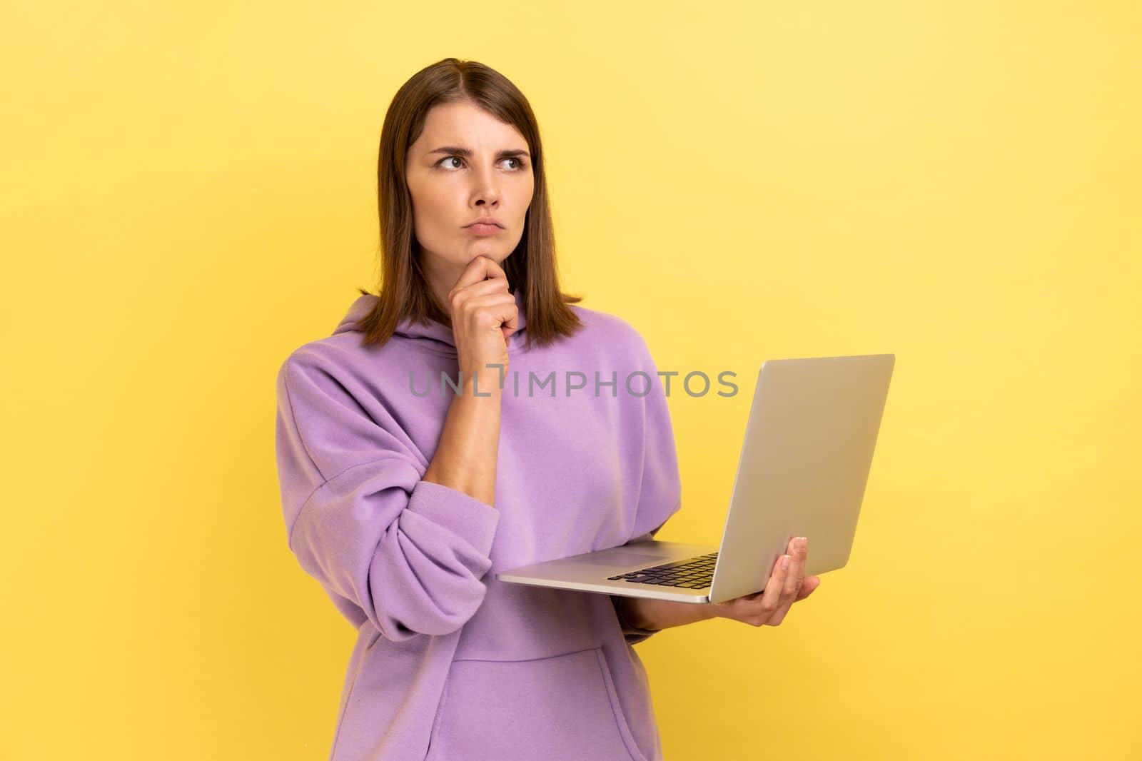 Pensive woman with working on laptop, holding chin, thinking about new project, looking away with thoughtful expression, wearing purple hoodie. Indoor studio shot isolated on yellow background.