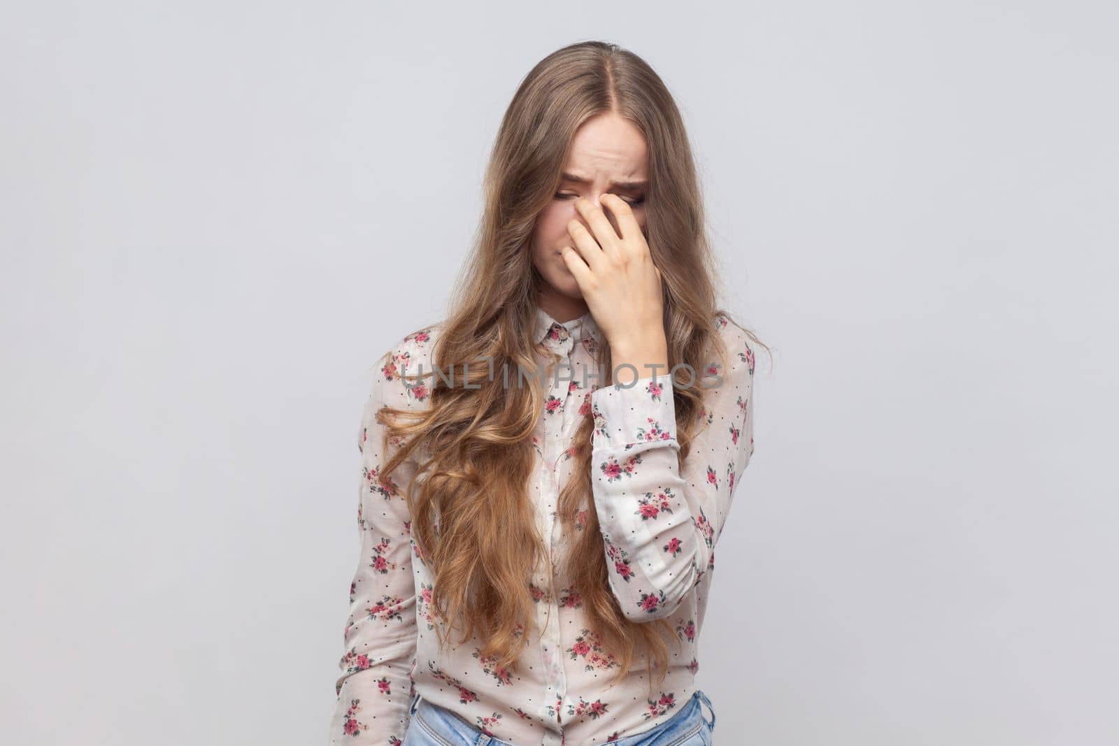 Portrait of desperate depressed woman with wavy blond hair rubbing eyes, crying because of hopelessness and loneliness, nervous breakdown. Indoor studio shot isolated on gray background.