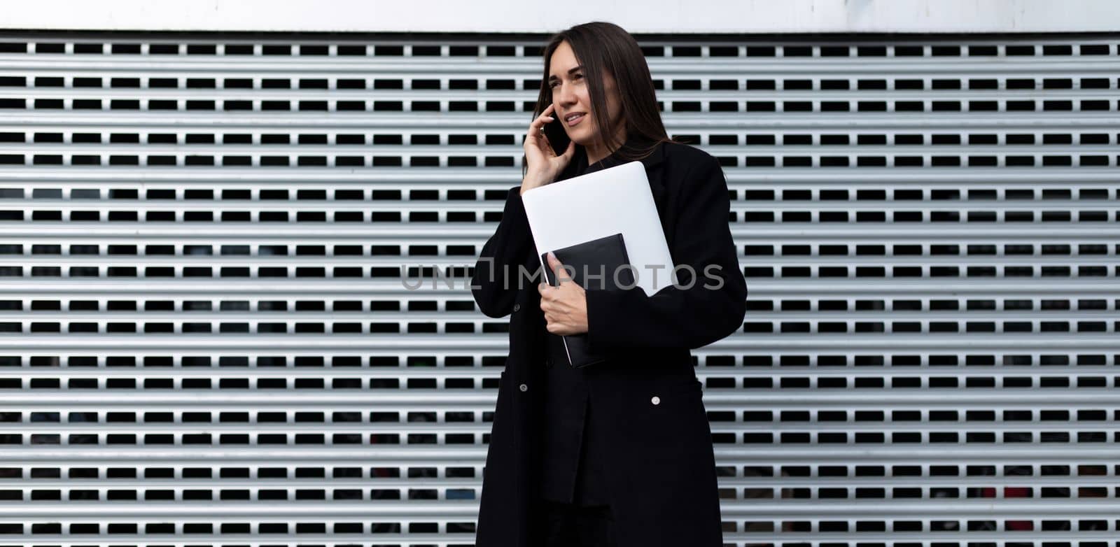 successful business woman with a laptop in her hands speaks on the phone against a gray wall by TRMK