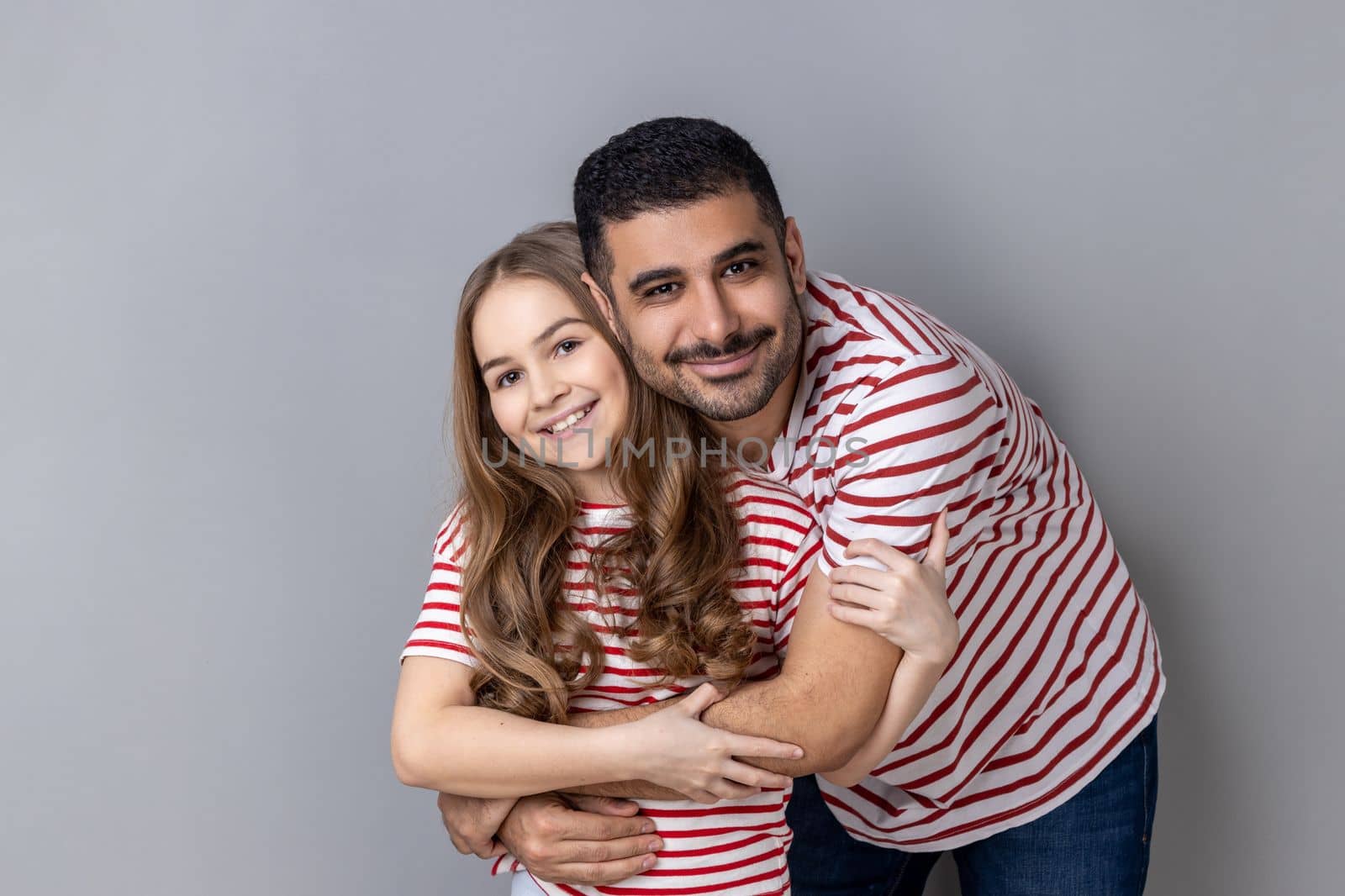 Portrait of loving happy father and daughter in striped T-shirts standing together, dad hugging cute little girl, looking at camera with smile. Indoor studio shot isolated on gray background.