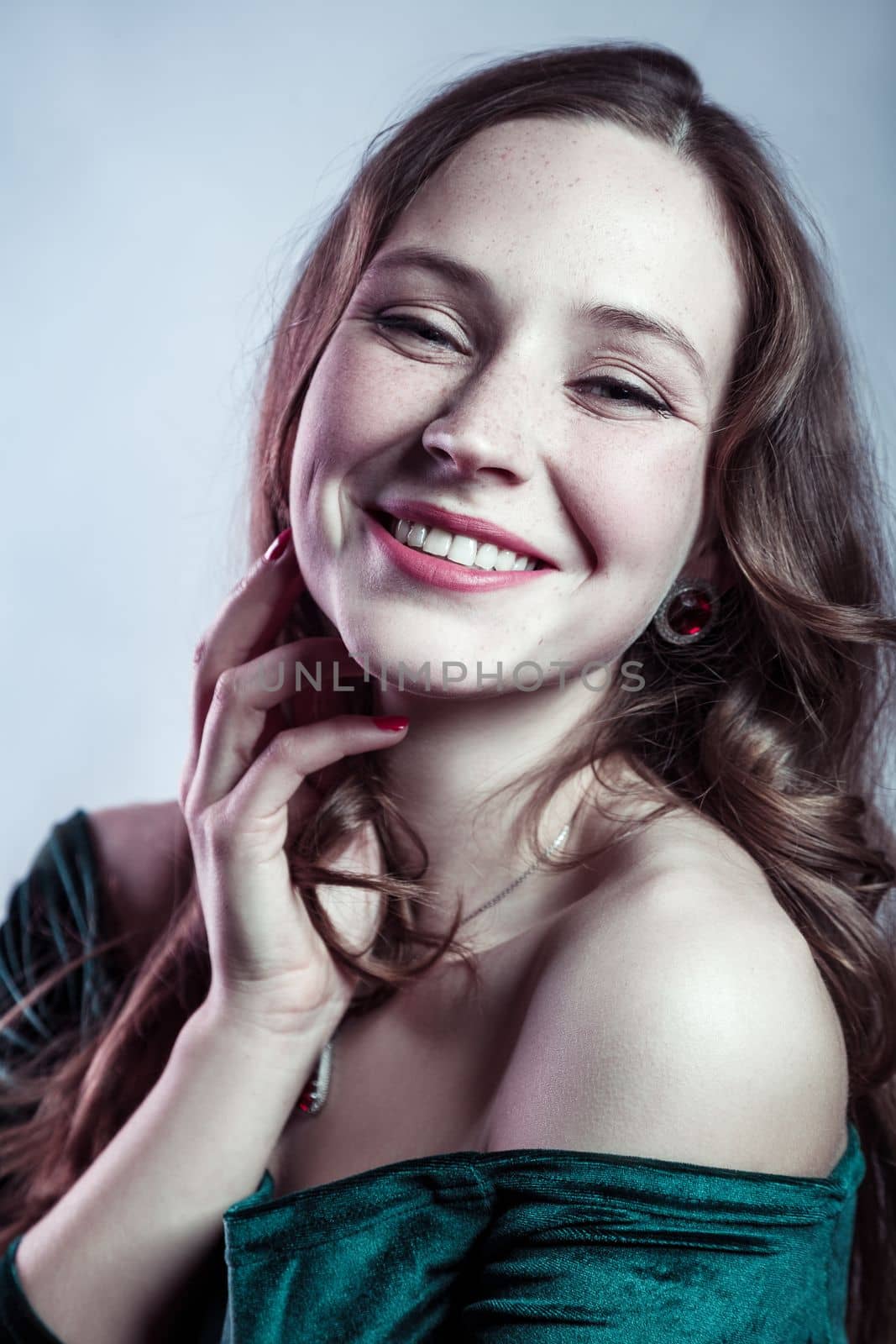 Beauty portrait of smiling positive satisfied elegant woman with wavy hair wearing green dress looking at camera with toothy smile. Indoor studio shot isolated on gray background.