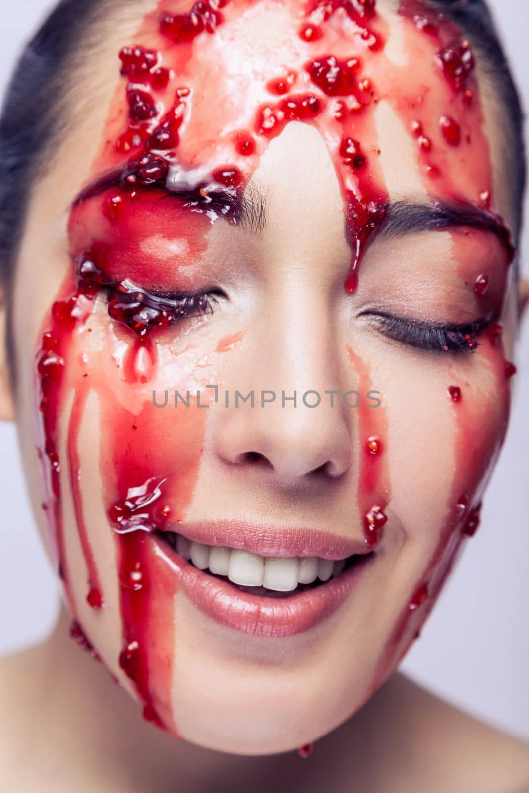 Closeup portrait of attractive woman fashion model with happy expression, smiling, keeps eyes closed, red jam flowing down on her face. Indoor studio shot isolated on gray background.